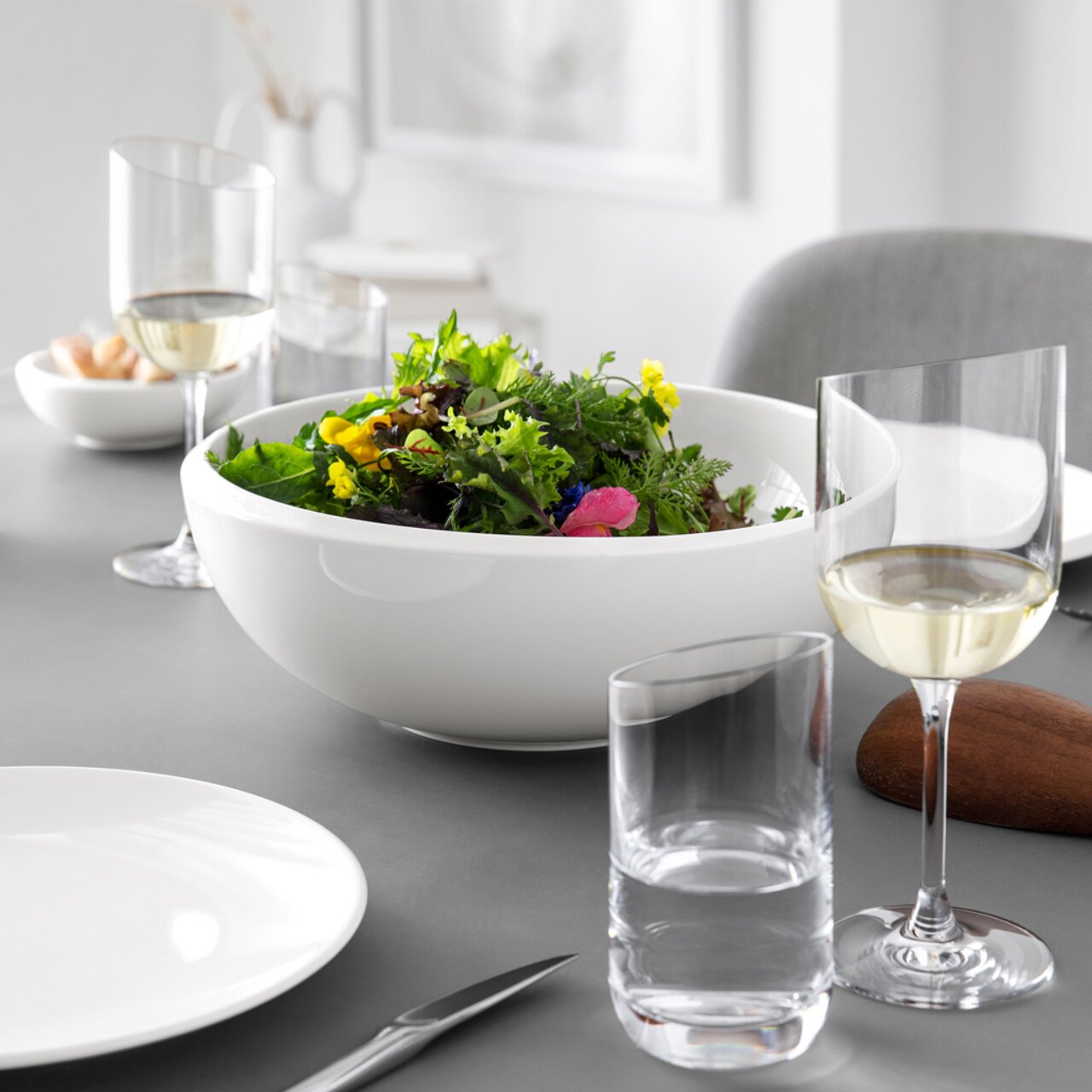 Elegante Tischszene mit einem großen weißen Salatschüssel im Vordergrund, umgeben von Weingläsern und Tellern, aus seitlicher Perspektive aufgenommen.