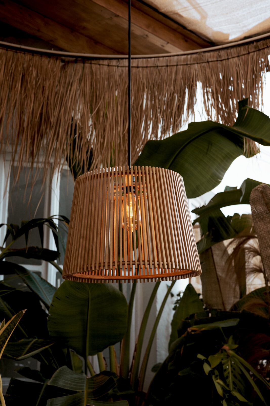 Hanglamp met een cilindervormige lampenkap van lichte houten lamellen, een warm lichtgevende gloeilamp en een zwarte kabel, gefotografeerd in een licht onderaanzicht tegen een achtergrond van tropische planten en een rieten dak