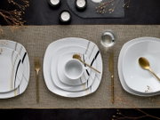 Draufsicht auf ein modern gedecktes Tisch-Arrangement mit weißem quadratischem Porzellan-Geschirr mit schwarzen und goldenen Linien, goldfarbenem Besteck, Tasse und Wasserglas auf beigem Leinenläufer