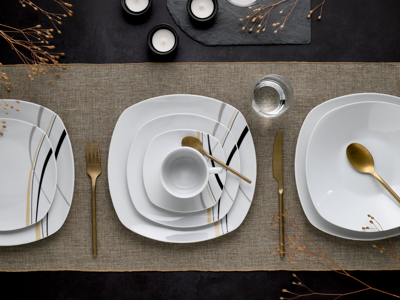 Draufsicht auf ein modern gedecktes Tisch-Arrangement mit weißem quadratischem Porzellan-Geschirr mit schwarzen und goldenen Linien, goldfarbenem Besteck, Tasse und Wasserglas auf beigem Leinenläufer