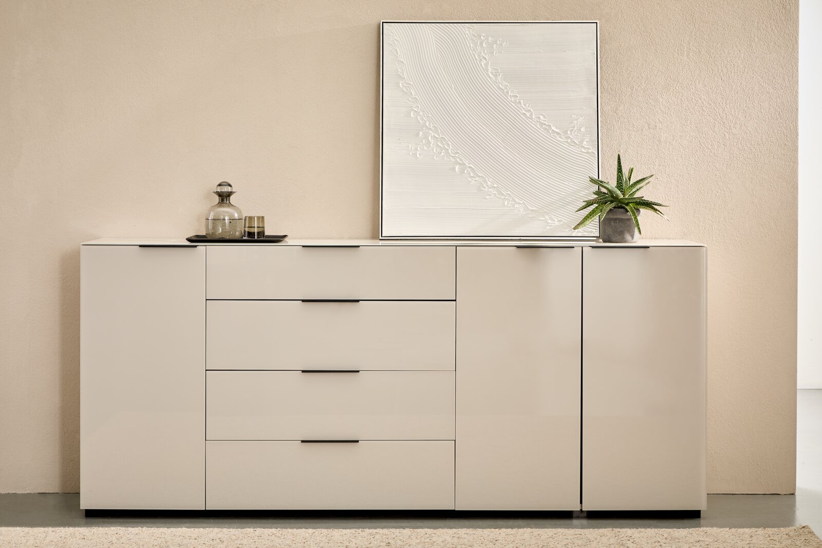 Novel XL-Sideboard MALIN 2.0 Modernes, weißes Sideboard mit drei Schubladen und zwei Türen, schwarze Griffleisten, dekoriert mit Glasflasche, Tablett, Pflanze und gerahmtem Reliefbild; frontale Ansicht auf Augenhöhe.