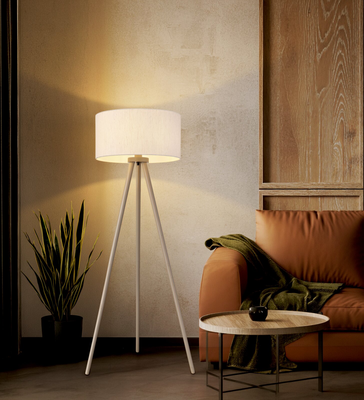 Weiße dreibeinige Stehlampe mit zylindrischem Stoffschirm, eingeschaltet im Wohnzimmer neben braunem Sessel und Beistelltisch, frontale Perspektive auf das Produkt