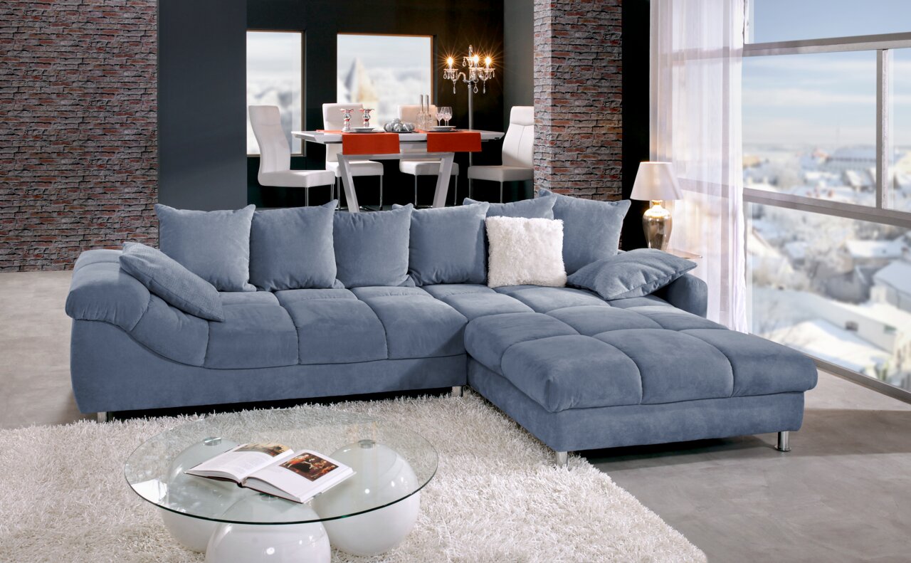 Z2 Ecksofa BROOKLYN II Blaue Rundecke (L) in einem modernen Wohnzimmer mit großen Fenstern, einem weißen Teppich und einem Glastisch, seitliche Perspektive.