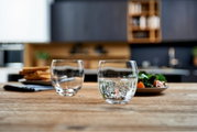 Zwei Trinkgläser Cheers 400ml auf einem Holztisch, gefüllt mit sprudelndem Wasser, im Vordergrund einer modernen Küche.