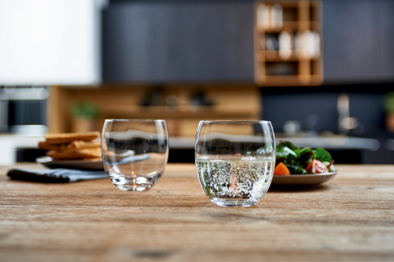 Zwei Trinkgläser Cheers 400ml auf einem Holztisch, gefüllt mit sprudelndem Wasser, im Vordergrund einer modernen Küche.