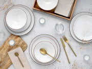 Draufsicht auf ein stilvolles Tischgedeck mit weiß gesprenkeltem Keramikgeschirr mit braunem Rand, goldfarbenem Besteck, Leinenservietten, Holztablett und kleinen Teelichtern auf heller Steinoberfläche