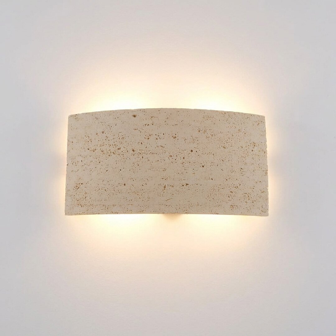 Beige wandlamp van natuursteen met een gebogen, rechthoekige vorm en warm indirect licht, van voren tegen de muur gefotografeerd.