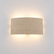 Beige wandlamp van natuursteen met een gebogen, rechthoekige vorm en warm indirect licht, van voren tegen de muur gefotografeerd.