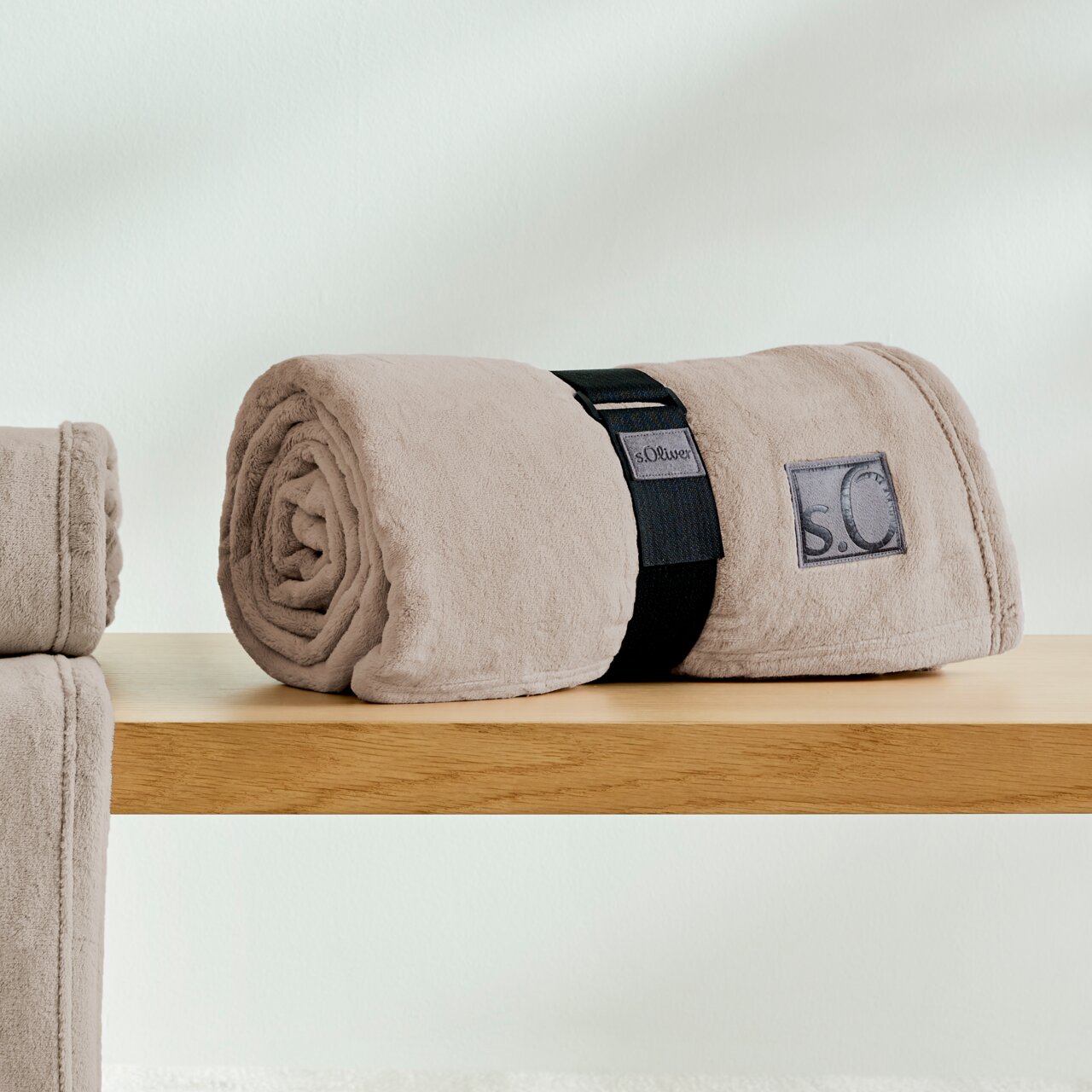 s.Oliver Decke Kuschelsoft Beige, gerollte Kuscheldecke von s.Oliver auf einem Holztisch, seitliche Perspektive