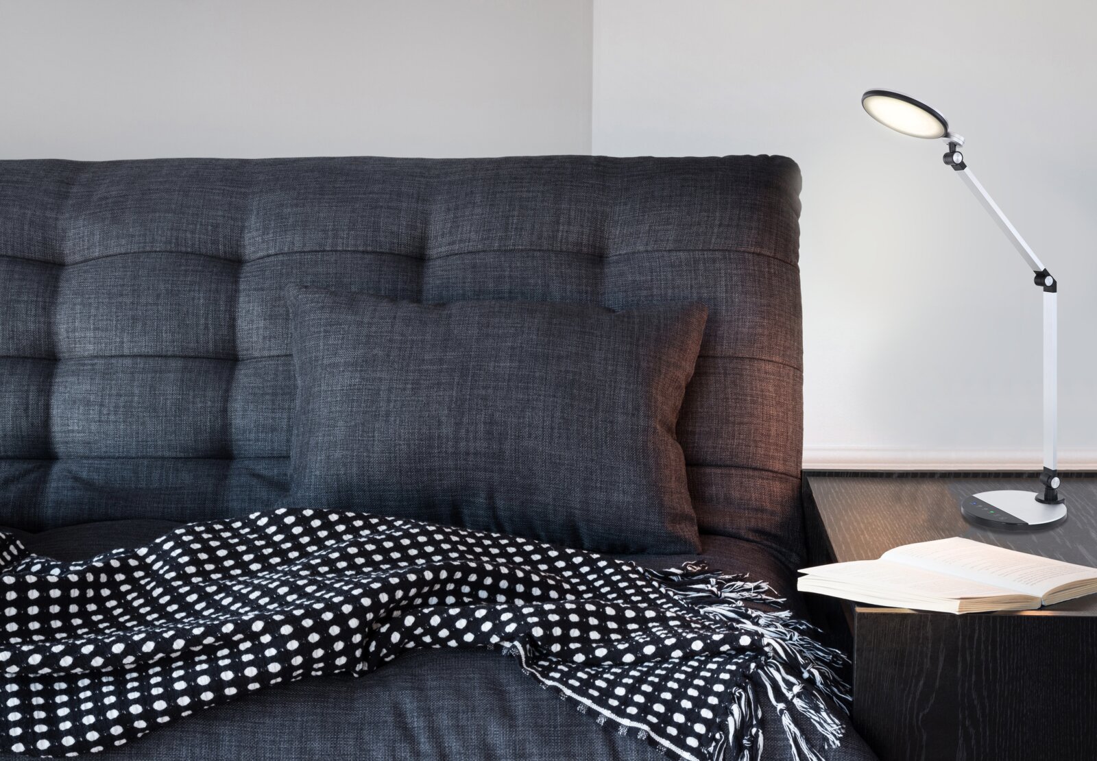 Moderne Tischleuchte auf einem Nachttisch neben einem Sofa mit grauem Kissen und schwarz-weiß gepunkteter Decke, seitliche Perspektive.