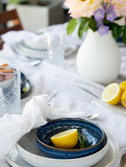 Gedeckter Esstisch mit blauer Keramikschale auf weißem Teller, Zitronenhälfte und Rosmarin, daneben Glas und weiße Vase mit Blumen, Perspektive: leicht von oben in Nahaufnahme.