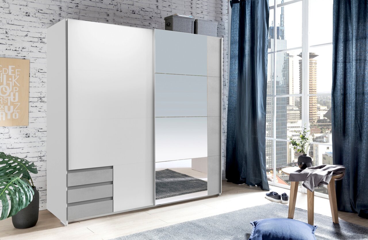 Moderner, zweiflügeliger Schwebetürenschrank in Weiß mit Spiegel, seitliche Perspektive, in einem hellen Raum mit großen Fenstern und urbanem Ausblick.