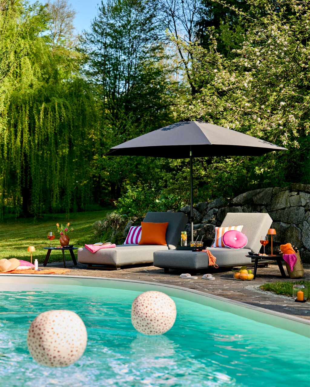 Amatio Gartenliege ENGADIN Pool-Lounge im Garten mit zwei grauen Sonnenliegen, bunten Kissen und schwarzem Sonnenschirm, fotografiert in leichter Untersicht aus Pool-Perspektive