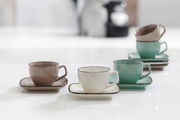 Drei Espressotassen mit Untertassen in verschiedenen Farben (grau, weiß, grün) auf einem Tisch, aus seitlicher Perspektive fotografiert.