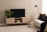 TV-Lowboard aus hellem Holz mit zwei Schubladen und offenen Fächern, darauf ein Fernseher, eine Pflanze und Kerzenhalter, seitliche Perspektive.