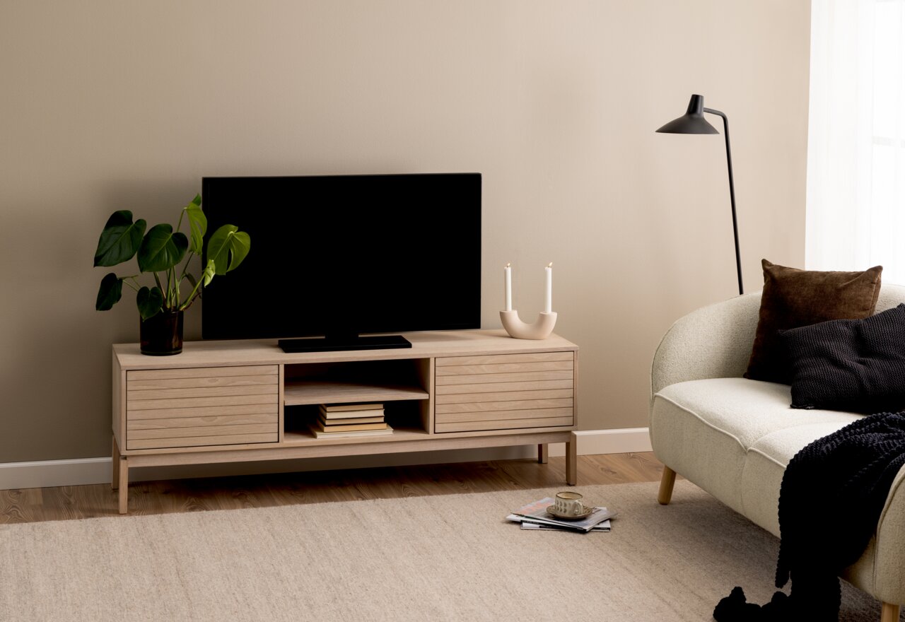 TV-Lowboard aus hellem Holz mit zwei Schubladen und offenen Fächern, darauf ein Fernseher, eine Pflanze und Kerzenhalter, seitliche Perspektive.