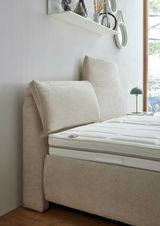 Seitliche Ansicht eines beigen Polsterbetts mit hohem Kopfteil und weißer Matratze, im Hintergrund ein Fenster und ein Regal mit Dekoration.