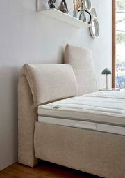 Seitliche Ansicht eines beigen Polsterbetts mit hohem Kopfteil und weißer Matratze, im Hintergrund ein Fenster und ein Regal mit Dekoration.