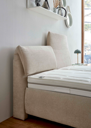 Seitliche Ansicht eines beigen Polsterbetts mit hohem Kopfteil und weißer Matratze, im Hintergrund ein Fenster und ein Regal mit Dekoration.