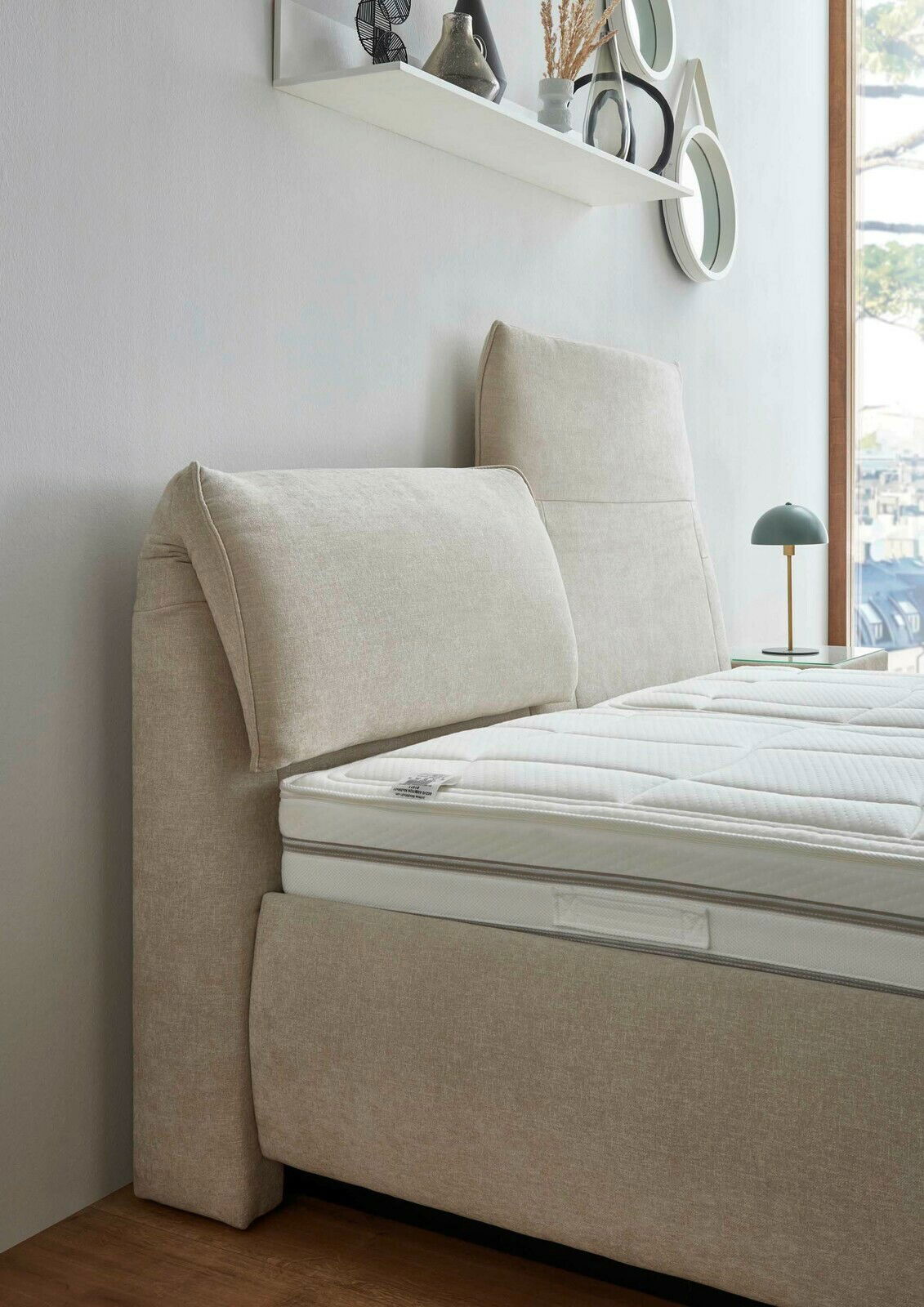 Seitliche Ansicht eines beigen Polsterbetts mit hohem Kopfteil und weißer Matratze, im Hintergrund ein Fenster und ein Regal mit Dekoration.