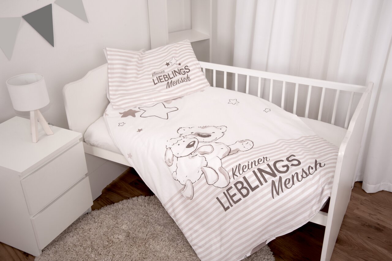 Kinderbett mit Renforcé-Bettwäsche-Set, bestehend aus einem Kissen und einer Decke mit niedlichem Bären- und Schafmotiv sowie dem Schriftzug 'Kleiner Lieblingsmensch', in einer hellen, gemütlichen Umgebung. Perspektive von schräg oben.