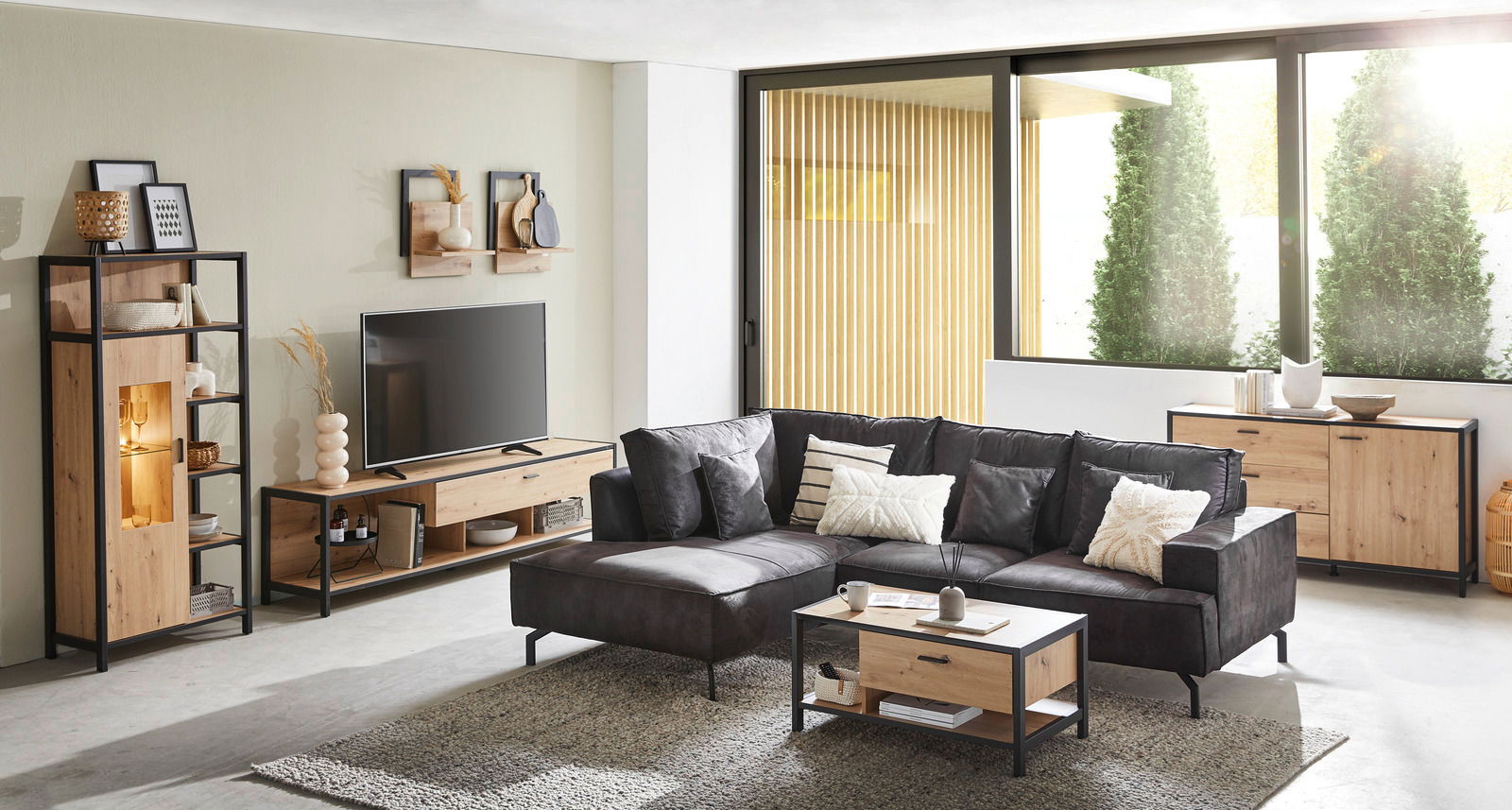 Moderne Wohnzimmermöbel in Holzoptik mit schwarzem Metallrahmen, bestehend aus TV-Lowboard, Vitrine, Couchtisch und Sideboard, aus der Perspektive eines hellen, geräumigen Wohnzimmers mit großen Fenstern.