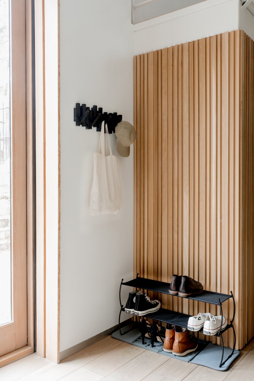 Moderne zwarte wandkapstok met haken, waaraan een tas en een hoed hangen, in een stijlvolle hal met houten lambrisering en een schoenenrek. Zijaanzicht.