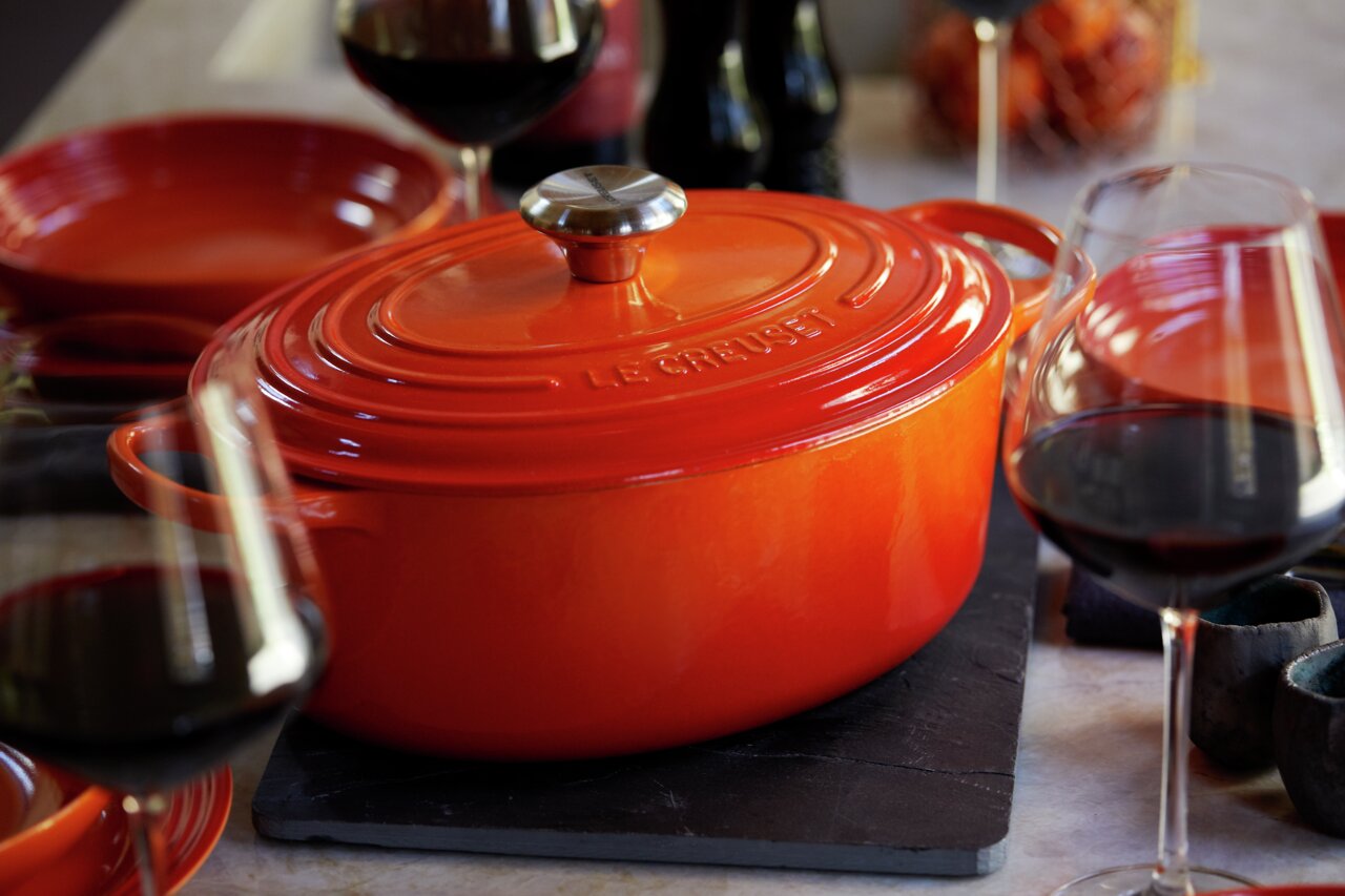 LE CREUSET Bräter oval 29 cm Ofenrot SIGNATURE Ofenroter Bräter von Le Creuset, 29 cm, auf einem Tisch mit Weingläsern, seitliche Perspektive