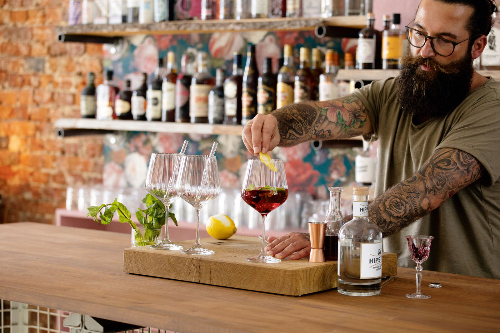 Ein Bartender mit tätowierten Armen bereitet einen Drink in einem stilvollen Ambiente zu. Auf einem Holztresen stehen mehrere Weingläser, eine Zitrone und eine Flasche Gin. Im Hintergrund sind Regale mit verschiedenen Spirituosen zu sehen. Perspektive: se