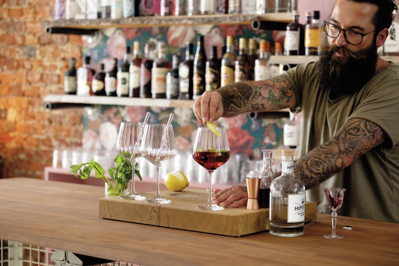 Een barman met getatoeëerde armen maakt een drankje klaar in een stijlvolle omgeving. Op een houten toog staan verschillende wijnglazen, een citroen en een fles gin. Op de achtergrond zijn planken met diverse sterke dranken te zien. Perspectief: se