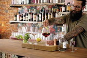 Een barman met getatoeëerde armen maakt een drankje klaar in een stijlvolle omgeving. Op een houten toog staan verschillende wijnglazen, een citroen en een fles gin. Op de achtergrond zijn planken met diverse sterke dranken te zien. Perspectief: se