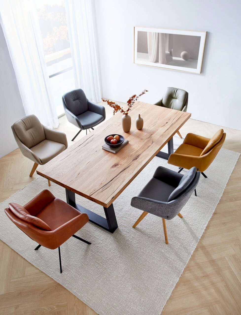 Waldwelt Armlehnstuhl ARUBA Esszimmer mit einem großen Holztisch und sechs verschiedenen Armlehnstühlen in einer modernen, hellen Umgebung, aus der Vogelperspektive betrachtet.