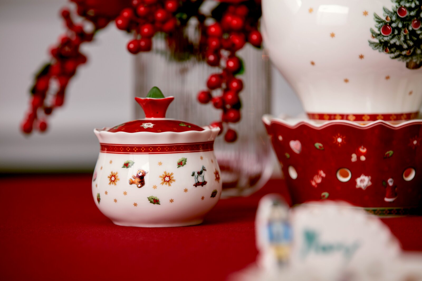 Weiß-rote Weihnachts-Zuckerdose aus Keramik mit Deckel und festlichen Motiven, frontal aus leichter Augenhöhe auf rotem Tischtuch präsentiert