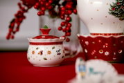 Weiß-rote Weihnachts-Zuckerdose aus Keramik mit Deckel und festlichen Motiven, frontal aus leichter Augenhöhe auf rotem Tischtuch präsentiert