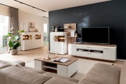 Modernes Wohnzimmer mit weißer Wohnwand und TV-Lowboard mit Holzoptik, Vitrine mit Glasfach und Beleuchtung sowie Couchtisch; Perspektive: leicht erhöhte Weitwinkel-Frontansicht schräg von links.