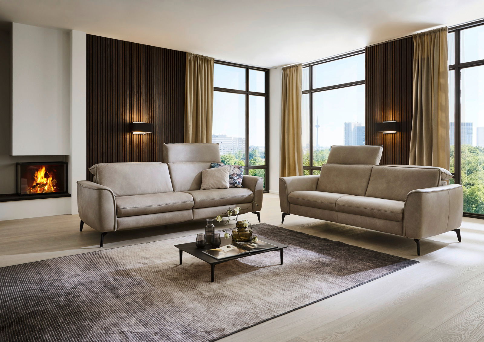 Modernes Wohnzimmer mit zwei beigen Sofas, eines davon ein 3-Sitzer und das andere ein 2,5-Sitzer, vor einer großen Fensterfront mit Blick auf die Stadt. Perspektive von vorne.