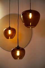 Drei hängende Leuchten mit bernsteinfarbenen Glaskörpern, die warmes Licht abstrahlen. Perspektive von unten, die die elegante Form und das sanfte Leuchten der Lampen betont.
