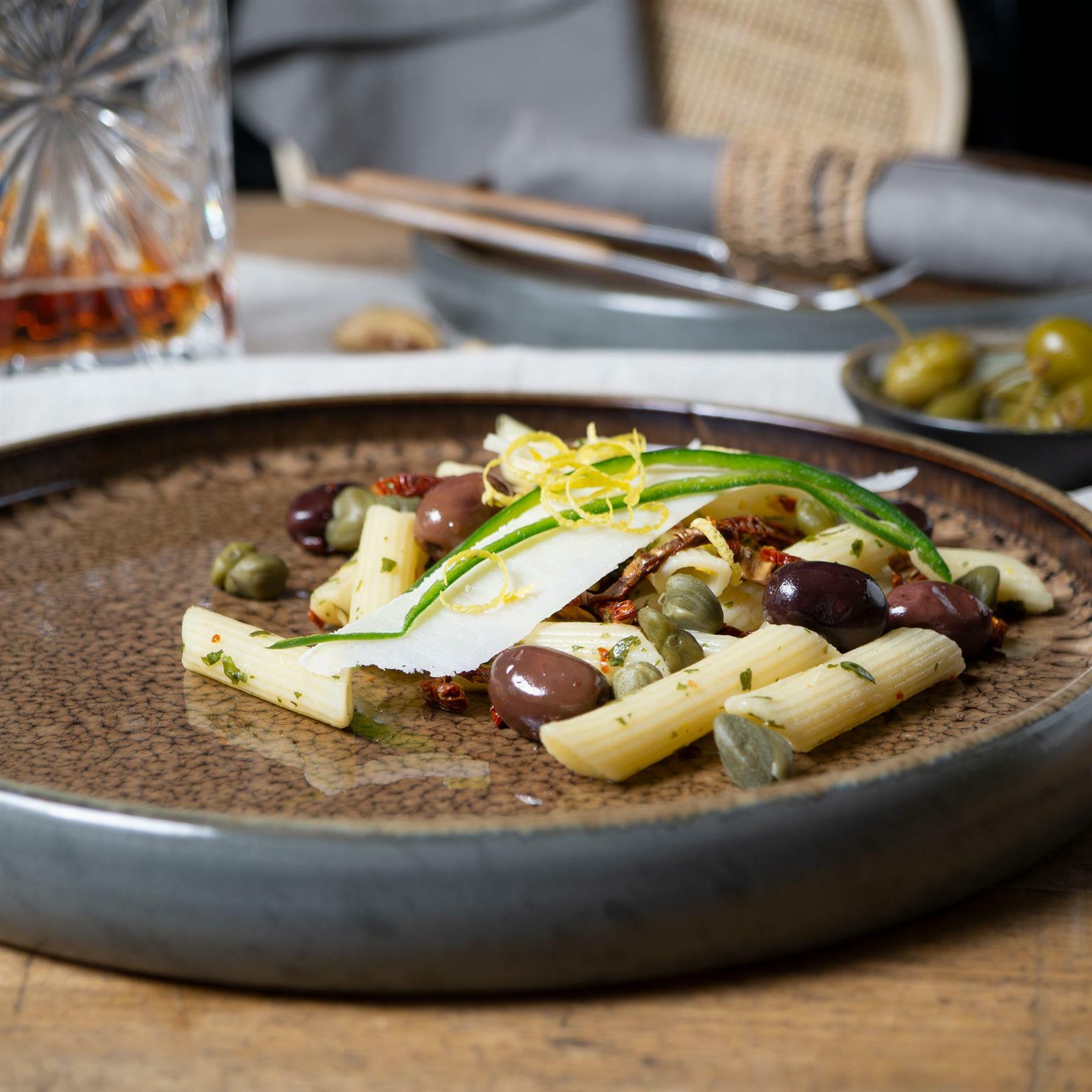 Nahaufnahme eines stilvoll angerichteten Tellers mit Pasta, Oliven und Kapern auf einem eleganten, beige-braunen Keramikteller.
