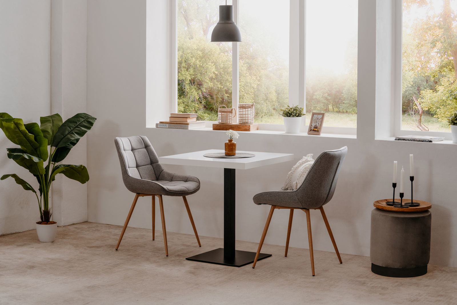 Z2 Esstisch QUADRATO Moderner Esstisch mit quadratischer weißer Tischplatte und schwarzem Metallfuß, umgeben von zwei grauen Polsterstühlen mit Holzbeinen, in einem hellen Raum mit großen Fenstern und Pflanzen, seitliche Perspektive.