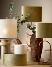 Verschillende lampenkappen in diverse maten en kleuren, waaronder een cilindervormige lampenkap in beige en goud, op een tafel gerangschikt. Het perspectief is frontaal.