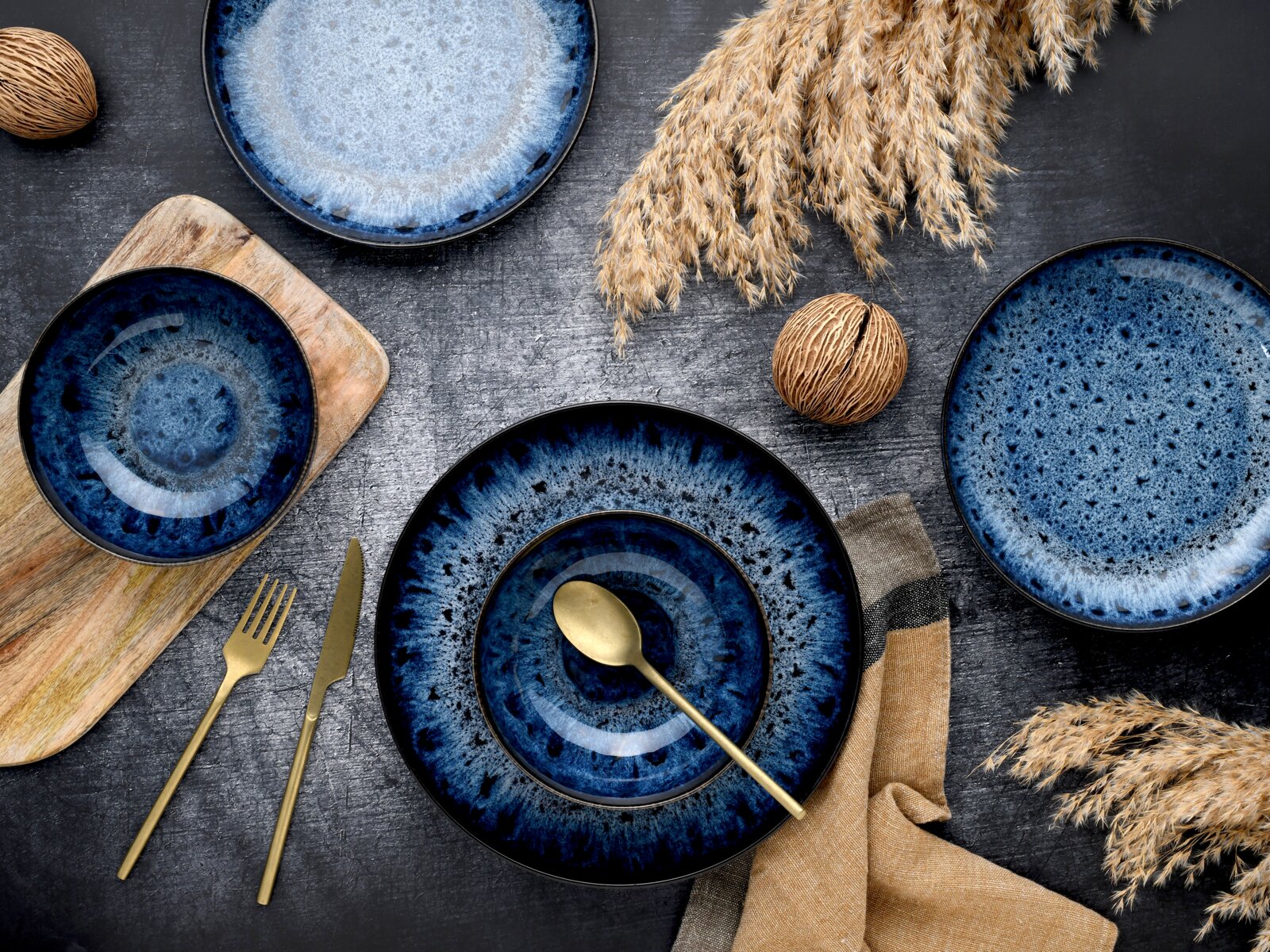 Dunkelblau gesprenkeltes Keramikgeschirr-Set mit Teller und Schalen, goldfarbenem Besteck und Stoffserviette auf dunklem Tisch, Draufsicht (Flatlay)
