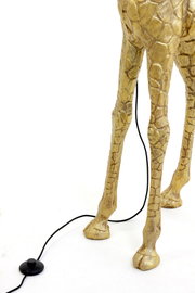 Close-up van de poten van een gouden staande lamp in de vorm van een giraffe, met een zwarte kabel en een voetschakelaar op een witte achtergrond.