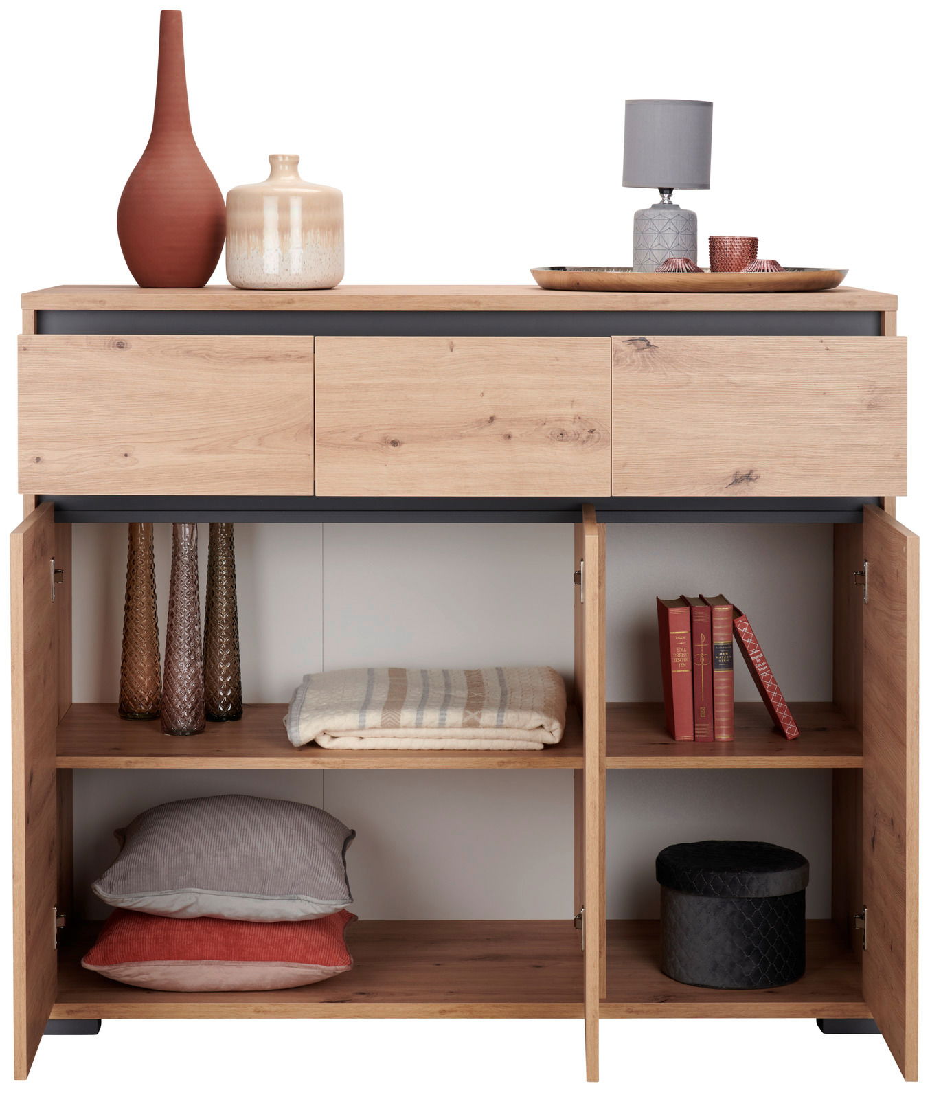 Sideboard aus Holz mit drei Schubladen und offenen Türen, gefüllt mit Dekorationsgegenständen und Textilien, aus der Frontperspektive.
