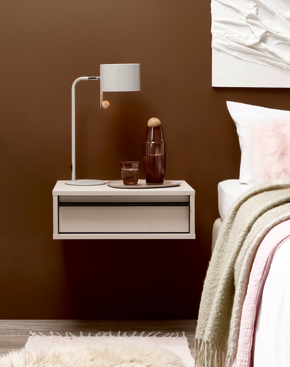 Z2 Nachtkommode PABLO Schwebender weißer Nachttisch mit Schublade neben einem Bett, darauf eine weiße Tischlampe sowie braune Glasflasche und Glas auf Tablett, frontal aus leicht seitlicher Perspektive vor brauner Wand