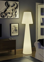 Leuchtende, moderne Stehlampe in Lampenform mit breitem Schirm und konischem Fuß in einem Wohnzimmer neben Sofa und Wandbildern, frontale Perspektive.