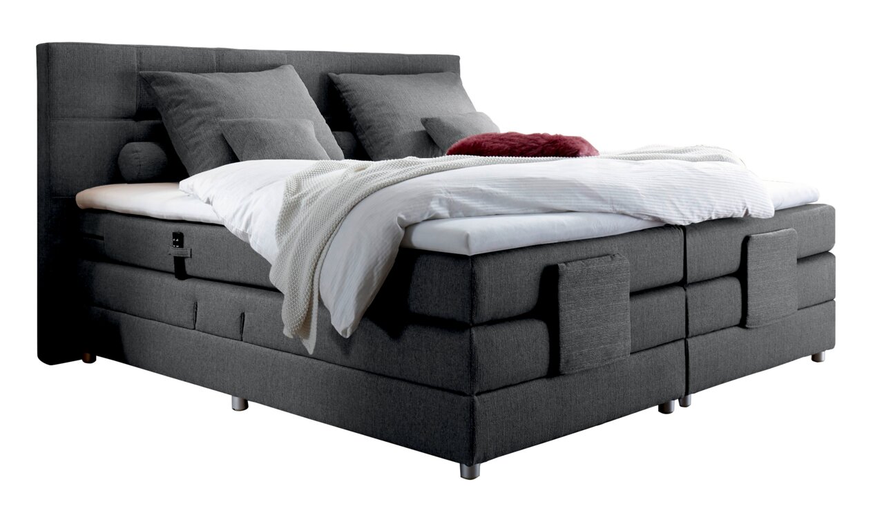 Graues Boxspringbett mit Kopfteil, seitliche Perspektive, dekoriert mit Kissen und Decken.