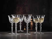 Sechs elegante Weißweinkelche aus Kristallglas mit feinem Schliffmuster, gefüllt mit Weißwein, auf einer Tischoberfläche vor dunklem Hintergrund, seitliche Perspektive.