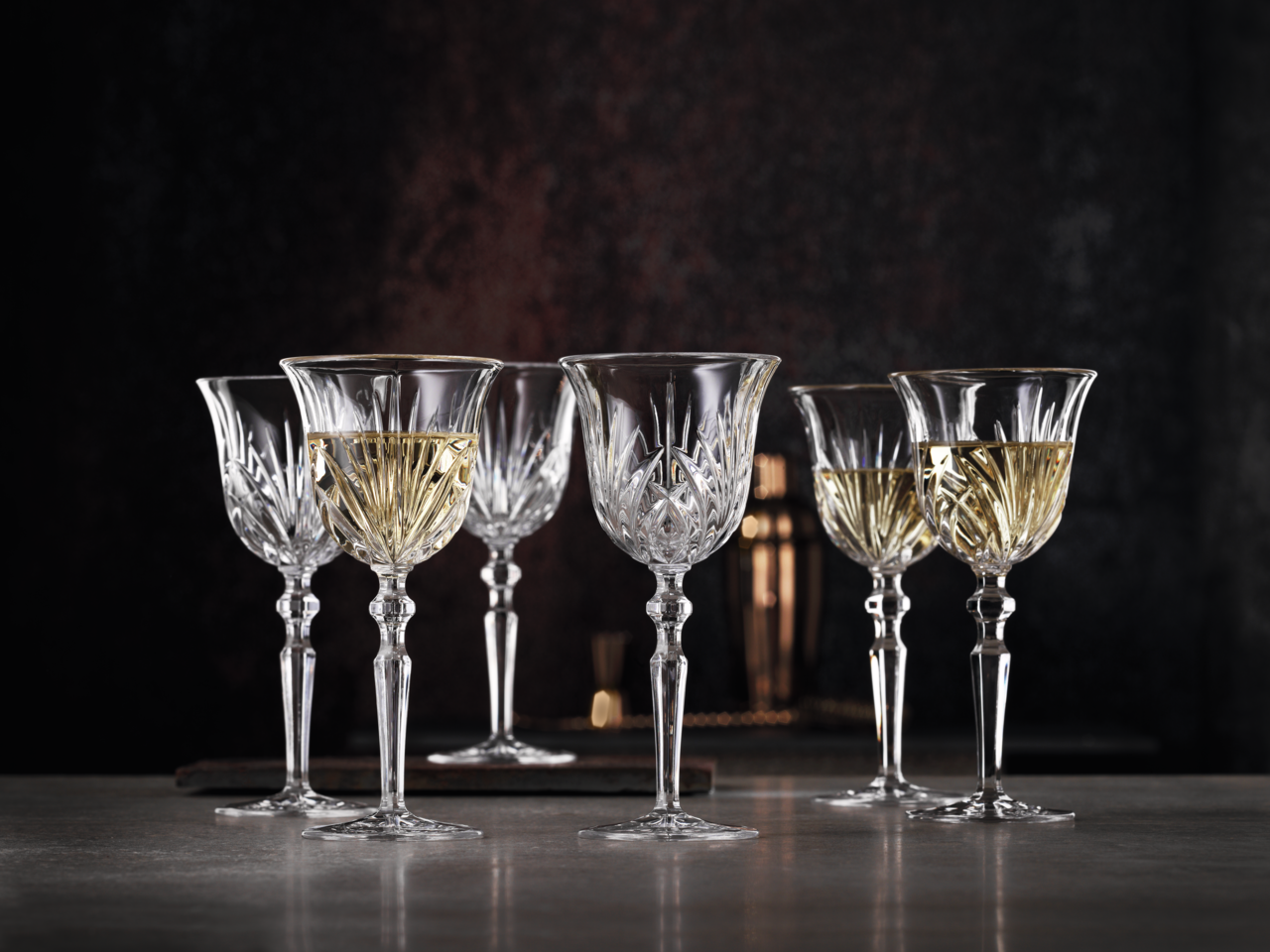 Sechs elegante Weißweinkelche aus Kristallglas mit feinem Schliffmuster, gefüllt mit Weißwein, auf einer Tischoberfläche vor dunklem Hintergrund, seitliche Perspektive.