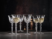 Sechs elegante Weißweinkelche aus Kristallglas mit feinem Schliffmuster, gefüllt mit Weißwein, auf einer Tischoberfläche vor dunklem Hintergrund, seitliche Perspektive.