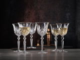 Sechs elegante Weißweinkelche aus Kristallglas mit feinem Schliffmuster, gefüllt mit Weißwein, auf einer Tischoberfläche vor dunklem Hintergrund, seitliche Perspektive.